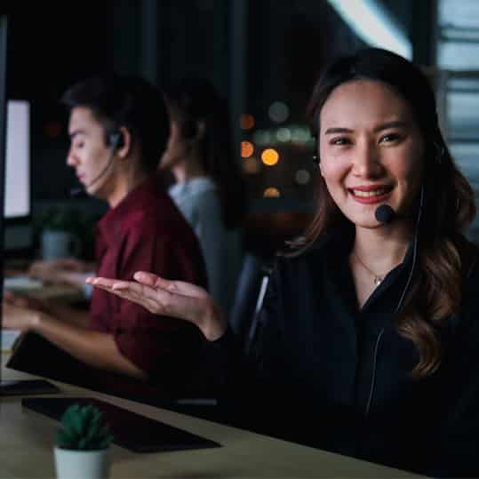 Telephone answering call center is busy picking up phone calls even though it is dark outside. There is a happy smiling young woman with headphones working at call center service desk with her teammates at night. Ready to take with customer on hands-free phone.