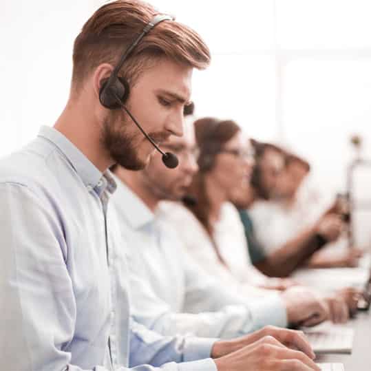 Man with a headset is talking to a patient. In the background, you can see multiple live answering service agents busy with callers.