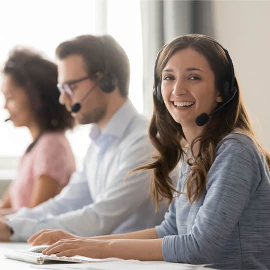 A happy woman is answering phone calls through her headset as she smiles at the camera. Her colleagues are busy answering phones behind her.