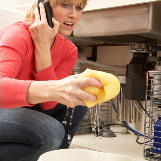 A Woman Mopping Up a Leaking Sink with a sponge is On the Phone trying to get a hold of a plumbing company or a plumber.